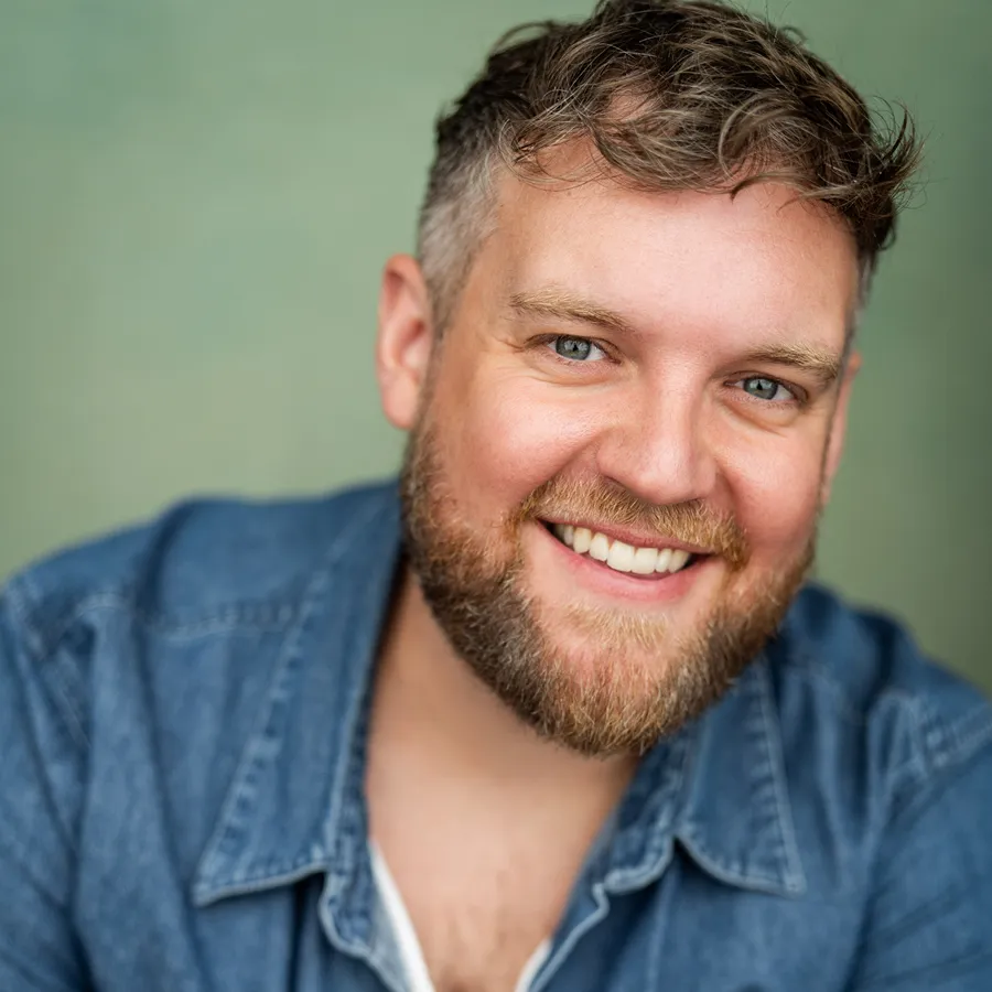 Dan Buckley, cast of One Day the Musical. Smiling headshot in a denim shirt.