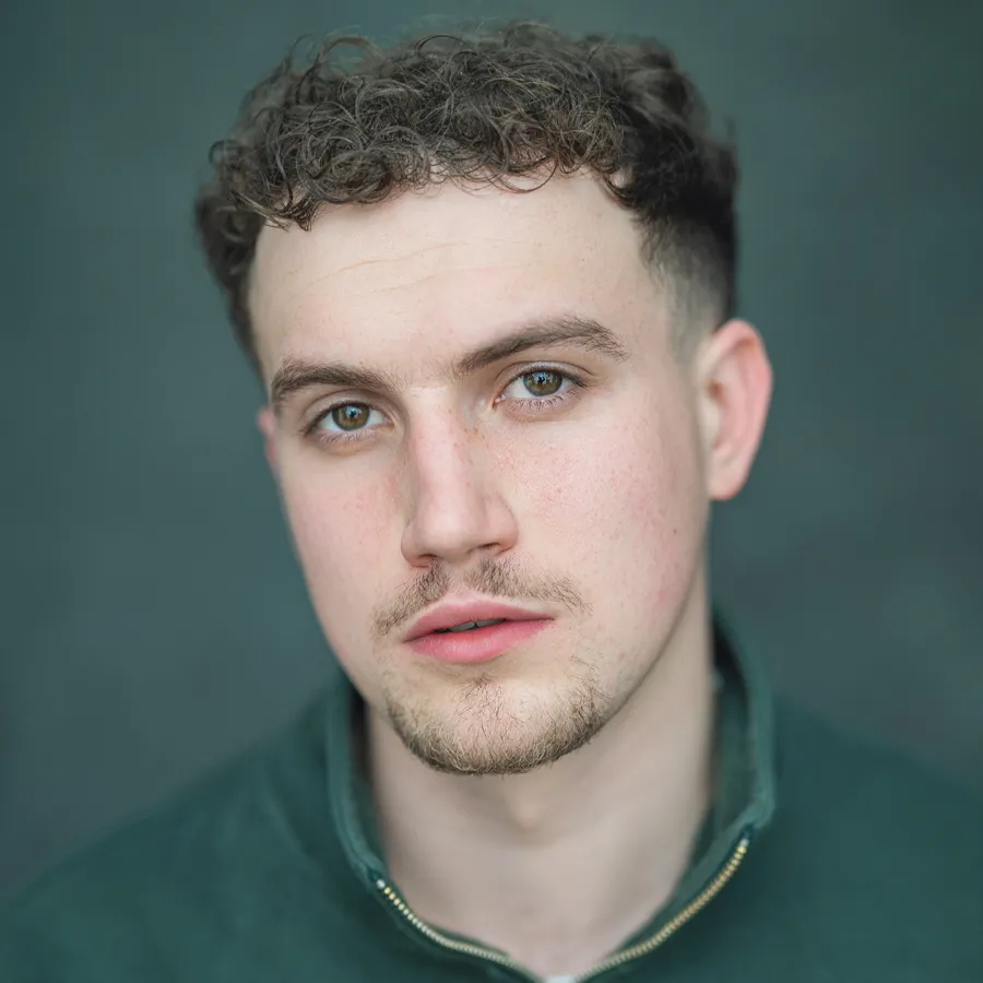 Ewan Murphy, cast of One Day the Musical. Headshot with curly hair, wearing a green zip-neck top.