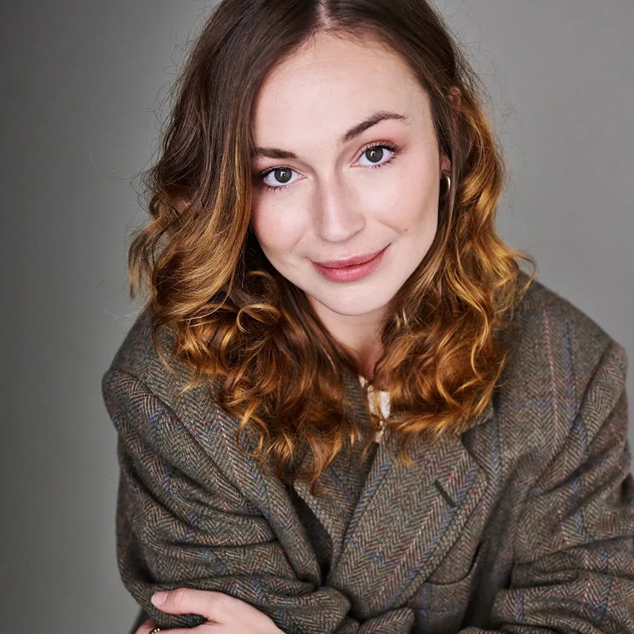 Jennifer Louise Jones, cast of One Day the Musical. Smiling headshot with wavy hair, wearing a tweed jacket.