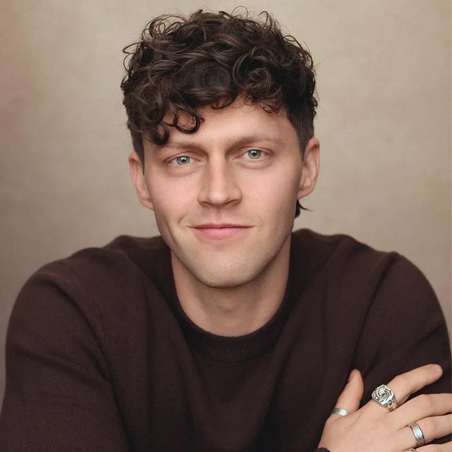 Tom Mussell, cast of One Day the Musical. Headshot with curly hair and arms folded, wearing a brown jumper.